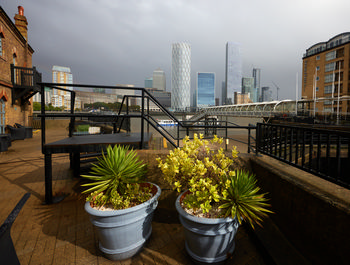 Canary wharf doubletree plants This urban photograph was taken during the afternoon in early autumn and shows potted plants in the outdoor area of the DoubleTree by Hilton in Canary Wharf, London, United Kingdom. The image foreground features two prominent blue-grey planters containing spiky evergreen and leafy shrubs, emphasizing decorative plants against the paved terrace. In the background, the iconic skyline of Canary Wharf rises, including prominent London landmarks such as One Canada Square and the cylindrical Landmark Pinnacle, all situated on the north bank of the River Thames. The photograph captures the modern architecture and high-rise buildings of this renowned London financial district, while overcast skies hint at typical autumn weather in the United Kingdom.